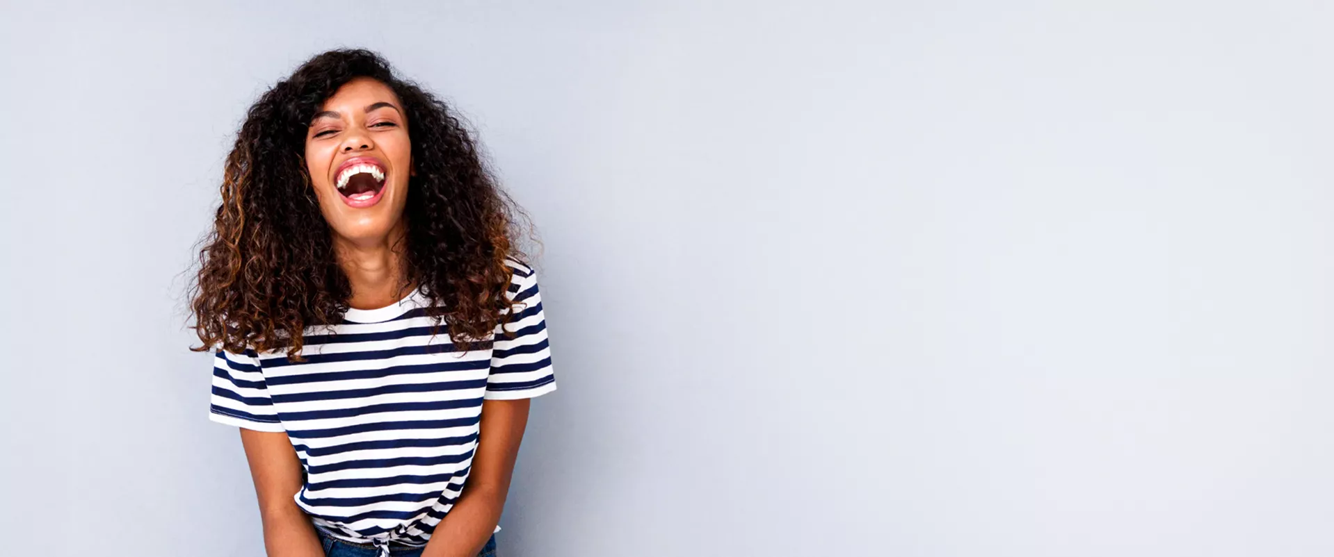 A smiling woman with curly hair wearing a navy-and-white striped shirt, standing against a plain light grey background