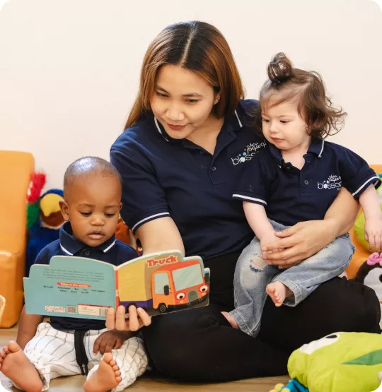 A nursery teacher in a navy Blossom Nursery & Preschool shirt sits on the floor reading a truck-themed picture book to two young children, who sit close beside her in a bright and playful classroom